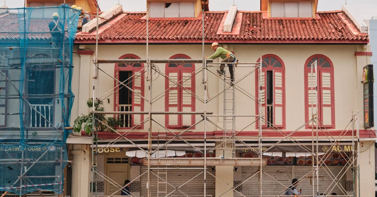Des ouvriers sur un échafaudage rénovent la façade d'un bâtiment historique avec un toit rouge et de hautes fenêtres.